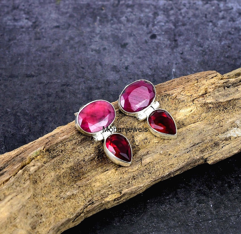 May include: A pair of silver earrings with oval and pear-shaped red gemstones. The earrings are displayed on a piece of wood. The brand name "Mohanjewels" is visible in the image.