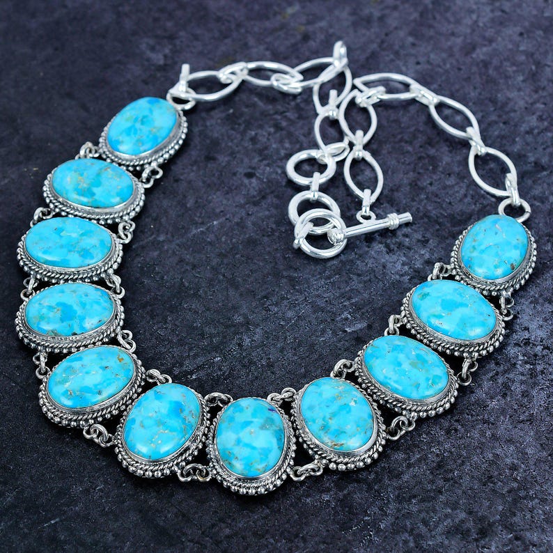 Op de afbeelding: Een zilveren ketting met ovale turkooizen stenen. De stenen zijn gezet in een decoratief zilveren frame en verbonden met een zilveren ketting. De ketting heeft een sleutelvormige sluiting. De turkooizen stenen hebben een helderblauwe kleur.
