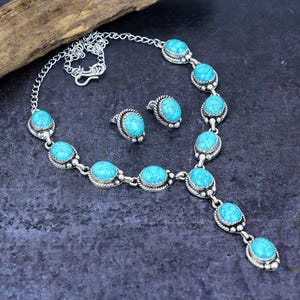 以下が含まれることがあります： 楕円形のターコイズストーンが特徴のシルバーネックレスとイヤリングのセット。ネックレスは、一連のターコイズストーンが付いたペンダントデザインです。イヤリングは小さなスタッドです。ジュエリーは暗い表面に表示されます。
