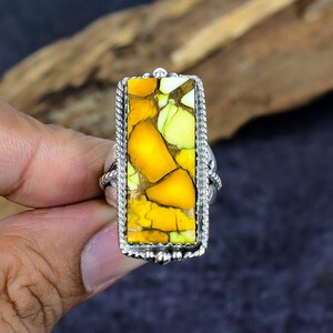 May include: Silver ring featuring a rectangular stone with a mosaic of yellow and green hues. The stone is framed by a twisted rope-like border and set on a silver band. The ring's design is eye-catching and suitable for everyday wear.