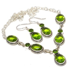 May include: A silver necklace and earring set with bright green, oval-cut gemstones. The necklace has a chain and multiple gemstone clusters. The earrings have a similar design, with a hook closure. The jewellery is set against a white background.