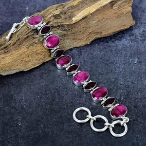 Puede incluir: Una pulsera de plata con piedras preciosas rosas oscuras de corte ovalado y marquesa. La pulsera está engastada en metal plateado y tiene un cierre de palanca. Las piedras están dispuestas en un patrón repetitivo.