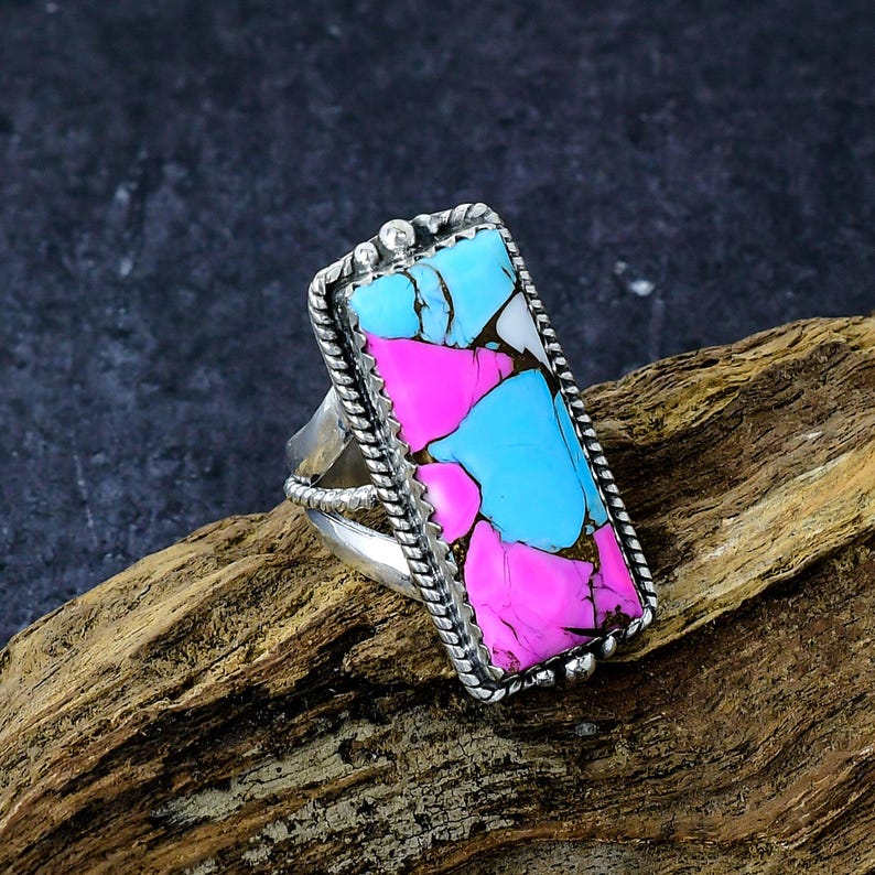 May include: A rectangular silver ring with a colorful stone inlay. The stone features a mosaic of pink, light blue, and white sections. The ring has a twisted rope design around the stone and a decorative band.
