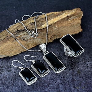 May include: A silver jewelry set featuring a necklace, earrings, ring, and pendant. Each piece has a rectangular black stone set in a silver frame with a beaded edge. The necklace has a delicate chain and a matching pendant.