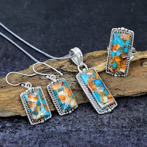 May include: A jewelry set featuring a pendant necklace, earrings, and a ring. Each piece has a rectangular turquoise, orange, and white stone set in a silver frame. The set is displayed on a wooden surface.