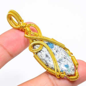 Può includere: Un ciondolo avvolto in filo d'oro con una pietra macchiata di blu e bianco. Il ciondolo è di forma ovale e ha un anello d'oro in alto per attaccarlo a una catena.