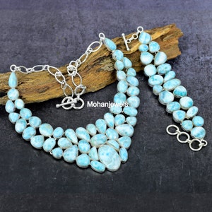 May include: A silver necklace and matching bracelet set featuring multiple polished, light blue, oval and teardrop-shaped Larimar stones set in silver bezels. The necklace has a toggle clasp and a silver chain, with the text Mohanjewels visible.