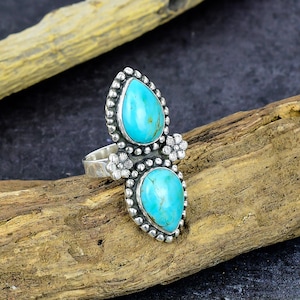 May include: A sterling silver ring featuring two teardrop-shaped turquoise stones set vertically. The stones are surrounded by a beaded metal border and accented with small silver flower details between them. The band is smooth and polished silver.