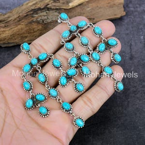 Puede incluir: Un collar de plata con múltiples hilos de piedras turquesas ovaladas. Las piedras están engastadas en un metal plateado con un borde con cuentas. El collar se muestra en una mano.