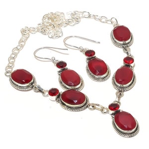 May include: A silver necklace and earrings set with red gemstone accents. The necklace features a chain with five oval-shaped red gemstones, each surrounded by a silver frame. The earrings are drop earrings with a single red gemstone.