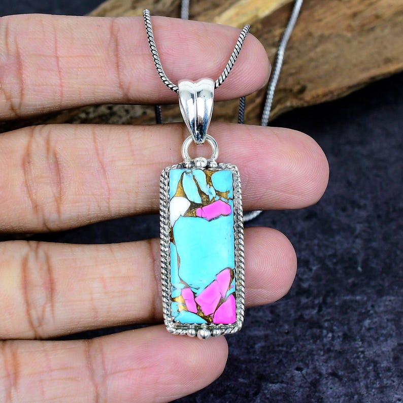 May include: A rectangular pendant necklace with a silver chain. The pendant features a turquoise, pink, white, and gold stone set in a silver frame. The pendant is approximately 1.5 inches long.