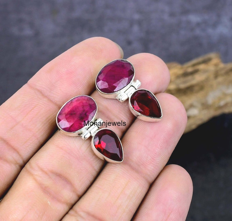 May include: Two pairs of gemstone earrings. Each earring features a large oval ruby and a smaller, teardrop-shaped garnet. The gemstones are set in silver. The text "mohanjewels" is visible.