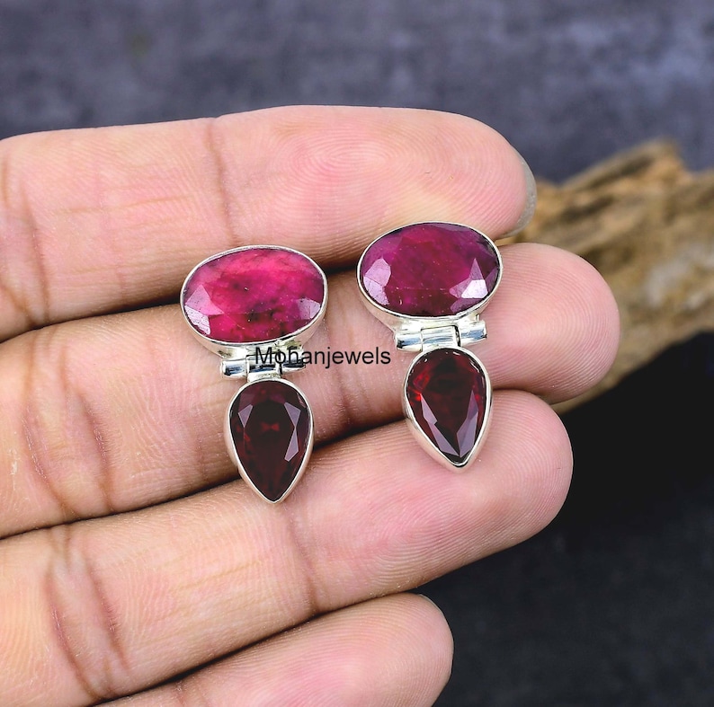 May include: A pair of sterling silver earrings featuring oval and teardrop-shaped red gemstones. The earrings have a polished finish and are set against a neutral background. The text "Mohanjewels" is visible on the image.