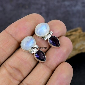 Puede incluir: Pendientes de plata con una piedra lunar y una amatista. La piedra lunar es ovalada e iridiscente, la amatista tiene forma de lágrima de color púrpura oscuro. Los pendientes están engarzados en plata. El texto 'Mohanjewels' es visible.