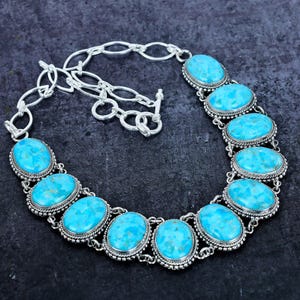 Op de afbeelding: Een zilverkleurige ketting met ovale turkooizen stenen. De stenen zijn gezet in een kralenrand en verbonden door een ketting. De ketting heeft een toggle sluiting. Donkere achtergrond.