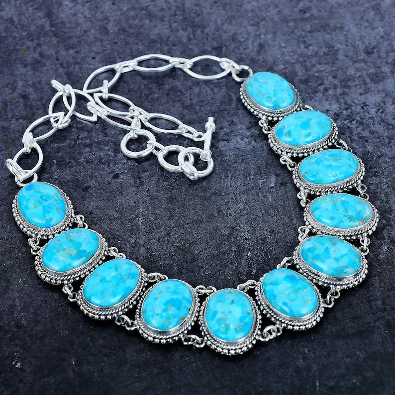 Op de afbeelding: Een zilverkleurige ketting met ovale turkooizen stenen. De stenen zijn gezet in een kralenrand en verbonden door een ketting. De ketting heeft een toggle sluiting. Donkere achtergrond.