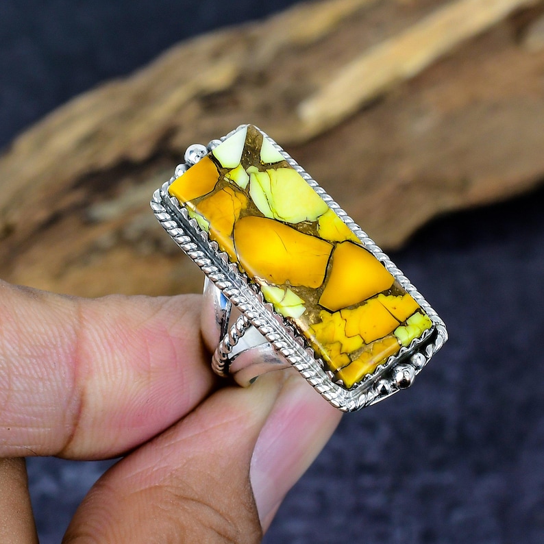 May include: A rectangular ring featuring a large, vibrant stone with yellow, green, and brown hues. The stone is set in a silver-colored metal with a textured rope-like border. The ring is held by a hand, showcasing the jewelry's design and color.