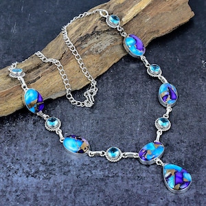 May include: A silver necklace featuring oval and teardrop-shaped turquoise and purple stones with gold accents. The necklace also includes small, round, light blue stones. The necklace is displayed on a piece of wood.