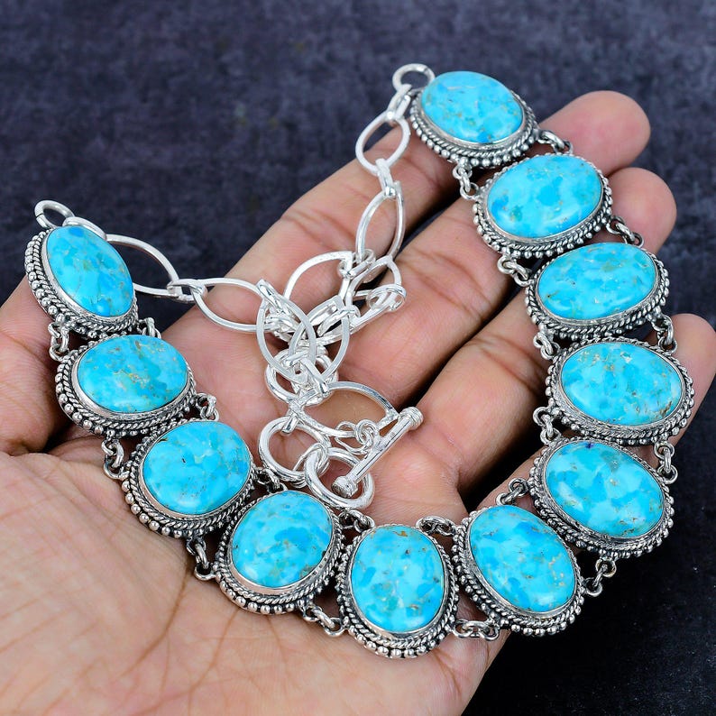 Op de afbeelding: Een zilveren ketting met ovale turkooizen stenen. De stenen zijn gezet in een gedetailleerd zilveren frame en verbonden door zilveren schakels. De ketting heeft een toggle sluiting.