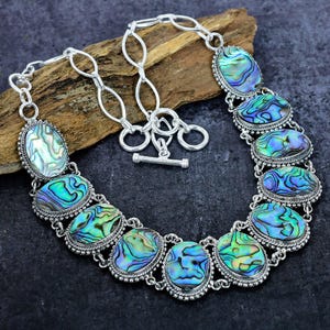 May include: A silver necklace featuring oval-shaped abalone shell pendants with iridescent blue, green, and purple hues. The pendants are set in a silver frame and connected by a silver chain with a toggle clasp. The necklace is displayed on a wooden surface.