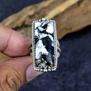 Puede incluir: Anillo rectangular de plata con una piedra llamativa con un patr&oacute;n en blanco y negro. La piedra est&aacute; engastada en un marco de plata detallado con un borde en forma de cuerda. Una joya audaz.