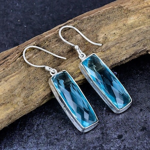 May include: A pair of silver earrings featuring rectangular, faceted, light blue gemstones. The earrings have a hook design and are displayed on a wooden surface against a dark background. The gemstones are set in a silver bezel.