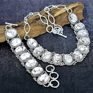 Könnte beinhalten: Ein Schmuckset mit Halskette und Armband. Die Schmuckstücke bestehen aus silberfarbenem Metall und haben ovale, weiße Steine mit braunen Adern. Die Halskette hat einen Knebelverschluss, das Armband einen Dreifachringverschluss.
