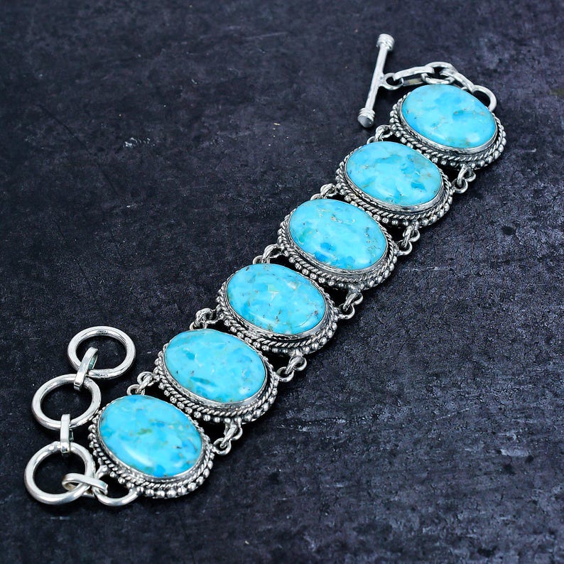 Op de afbeelding: Een zilveren armband met zes ovale turkooizen stenen. Elke steen is gezet in een gedetailleerde zilveren lijst met een parelrand. De armband heeft een tuimelsluiting en verbindingsschakels.