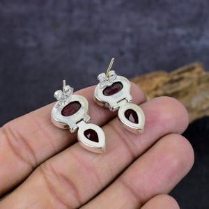 May include: A pair of silver-toned stud earrings with a teardrop-shaped red gemstone pendant. The earrings have a unique design with a small oval-shaped red stone at the top. The earrings are displayed on a neutral background.