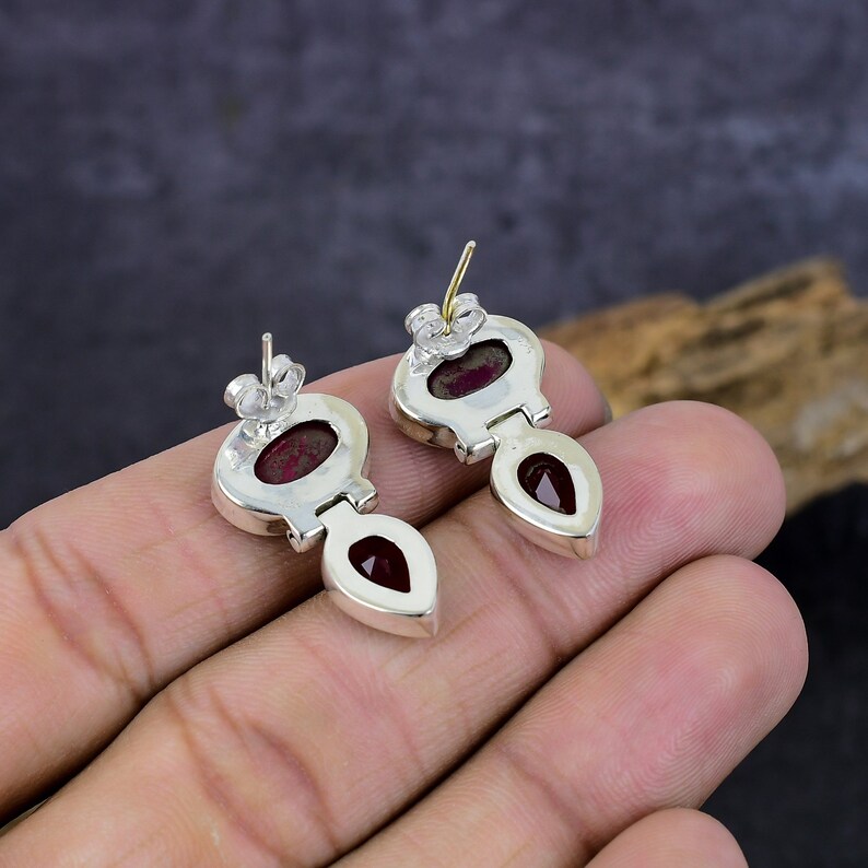 May include: A pair of silver-toned stud earrings with a teardrop-shaped red gemstone pendant. The earrings have a unique design with a small oval-shaped red stone at the top. The earrings are displayed on a neutral background.