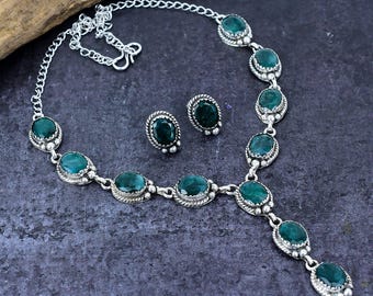 Puede incluir: Un conjunto de collar y pendientes de plata con piedras preciosas ovaladas de color verde esmeralda. El collar tiene una cadena y un colgante con varias piedras. Los pendientes son de tipo botón. Las joyas se exhiben sobre una superficie oscura.