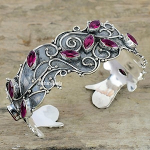 May include: A silver cuff bracelet featuring a floral design and pink gemstones. The bracelet is open on one side and has a hinged closure on the other.