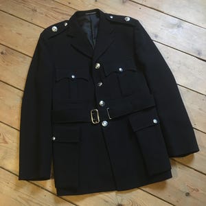 Puede incluir: Chaqueta negra con botones plateados y cinturón con hebilla dorada. La chaqueta tiene múltiples bolsillos y charreteras en los hombros. La chaqueta de tela oscura se exhibe sobre una superficie de madera.