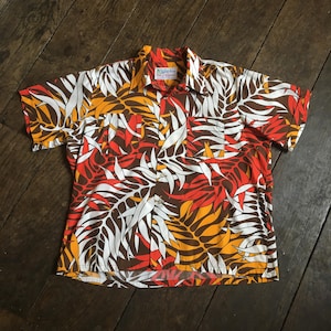 Peut inclure: Une chemise à manches courtes boutonnée avec un motif de feuilles tropicales aux tons orange, jaune, blanc et marron foncé. La chemise présente un col classique, une fermeture boutonnée sur le devant et deux poches de poitrine symétriques.