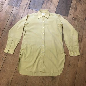May include: A long-sleeved, button-down shirt in a pale yellow color. The shirt has a classic collar, buttoned cuffs, and a curved hemline. The shirt is laid flat on a wooden surface.