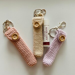 May include: Three crocheted keychains with wooden buttons and metal key rings. The keychains are in pink, beige, and purple. One keychain has a lip balm tube attached to it that says "ALOE LIPS" with "With Jojoba" printed below.