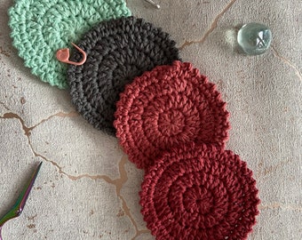 May include: Four crocheted coasters in different colors: green, gray, red, and burgundy. The coasters are arranged in a stack, with the green coaster on top and the burgundy coaster on the bottom.