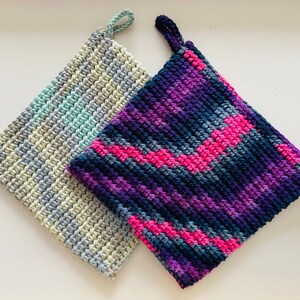 Puede incluir: Dos agarraderas de olla de ganchillo, una con un patrón a rayas de azul claro, gris y blanco, y la otra con un patrón multicolor de rosa, morado y azul.
