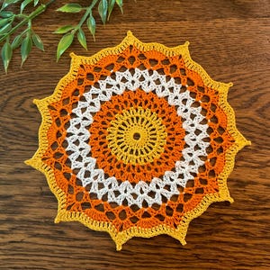 May include: A circular crochet doily featuring a star-like design with concentric rings of yellow, orange, and white yarn. The intricate needlework displays a central yellow core, followed by alternating bands of orange and white, ending in a yellow pointed edge.