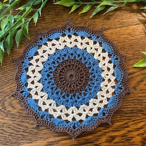 May include: A handmade, circular crochet coaster in shades of brown, blue, and cream. The coaster features a detailed, openwork design with a scalloped edge. The coaster is placed on a wooden surface, with green foliage in the background.