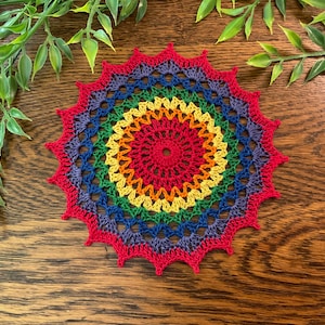 May include: A circular crochet doily featuring a vibrant rainbow color scheme. The design consists of concentric rings in red, orange, yellow, green, blue, and purple, finished with a pointed, star-like red border with intricate openwork stitching.