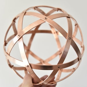 May include: A copper-toned metal sphere with an openwork design, constructed from intersecting bands secured with rivets. The bands form a geometric pattern. The sphere is held against a plain white backdrop.