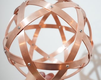 Op de afbeelding: Een koperkleurige bol bestaande uit elkaar kruisende metalen banden, vastgezet met kleine zilveren klinknagels. De banden creëren een open, geometrisch ontwerp. De bol wordt tegen een effen witte achtergrond gehouden.