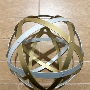 May include: A decorative metal sphere made of intersecting gold and silver bands. The bands are riveted together, creating a geometric design. This modern sculpture is a stylish accent piece for any home.