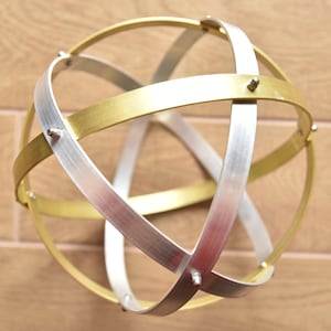 Può includere: Sfera decorativa composta da fasce metalliche incrociate, in tonalità oro e argento. Le fasce sono unite da rivetti, creando un motivo geometrico. Un elegante oggetto decorativo.
