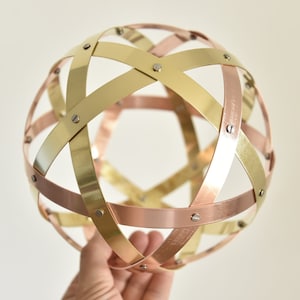 Può includere: Una sfera decorativa in metallo composta da fasce intersecanti nei toni dell'oro, del rame e dell'ottone. Le fasce sono fissate con piccole viti argentate, formando un design geometrico traforato. La sfera è tenuta in mano su uno sfondo neutro.