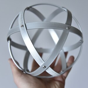 Può includere: Una sfera metallica color argento composta da fasce intersecanti, tenuta in mano. Le fasce sono fissate con piccoli rivetti, creando un design geometrico traforato. La sfera ha una finitura opaca e un'estetica moderna. Diametro: circa 10 cm.