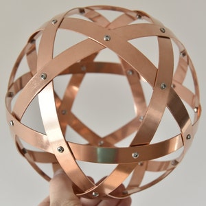 Può includere: Una scultura sferica in rame composta da fasce intersecanti, fissate con piccoli rivetti argentati. Le fasce creano un design geometrico aperto. La sfera è tenuta in mano, mostrando la sua forma tridimensionale. Il diametro è di circa 15 cm.