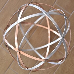 Può includere: Una scultura sferica composta da fasce metalliche intersecanti. Le fasce, alternate in argento e rame, sono fissate con piccoli rivetti. Il design geometrico crea un globo tridimensionale aperto, adatto all'arredamento domestico.
