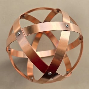 May include: A copper sphere sculpture composed of intersecting bands, secured with small silver rivets. The bands create a geometric pattern, reflecting light and showcasing the warm metallic color. This decorative piece could be used as a home decor accent.